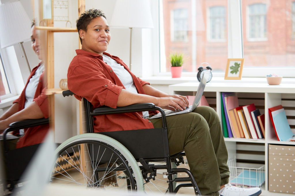 Portrait of African young woman with disabilities sitting in wheelchair and looking at camera while using laptop computer at home