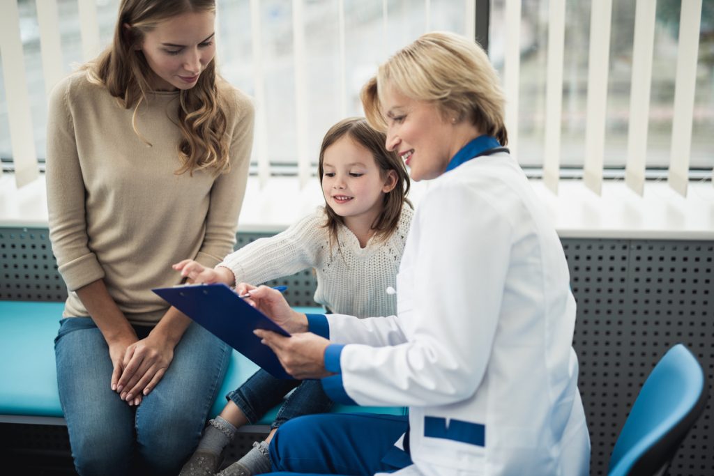 Concept of qualified consultation in healthcare system. Smiling little patient girl on couch with her mother studying notes of doctor in medical office